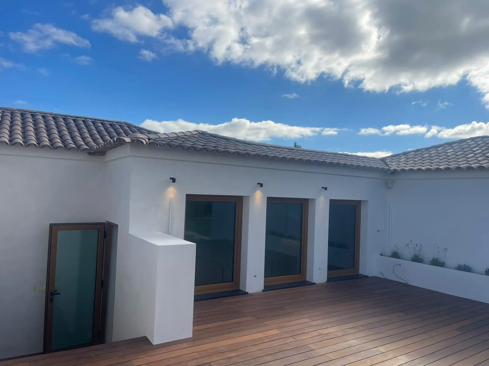 Terraço com deque de madeira e paredes brancas contínuas da moradia sob céu com nuvens